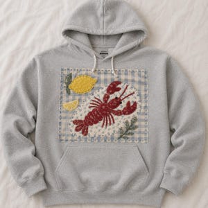 May include: A light gray hooded sweatshirt featuring a large rectangular patch on the front. The patch has a blue and white gingham border and displays a red lobster, a yellow lemon, and a lemon slice, all rendered in a floral fabric pattern.