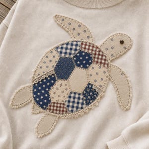 May include: A beige sweatshirt featuring a large turtle applique on the front. The turtle shell is constructed from various fabric patches in blue, white, and brown, including gingham, star, and floral patterns, accented with delicate lace trim.