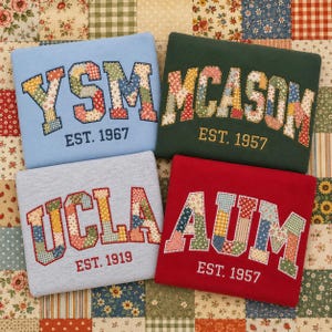 May include: Four folded sweatshirts in light blue, dark green, heather gray, and red. Each features large collegiate-style letters filled with colorful floral and gingham patchwork fabric, accompanied by embroidered text reading EST. 1967, 1957, 1919, or 1957.