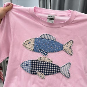 May include: A light pink short-sleeved t-shirt featuring two appliquéd fish designs. The top fish has a blue floral pattern, while the bottom fish has a navy and white gingham pattern. Both fish have visible blanket stitching and an "X" for an eye.