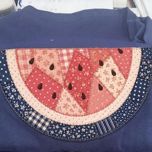 May include: A navy blue t-shirt featuring a large, semicircular watermelon slice applique made from various patchwork fabric scraps in shades of pink, red, and white, accented with dark brown teardrop-shaped seeds and a dark blue floral rind.