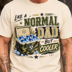 May include: Cream-colored t-shirt with the text "Like a Normal Dad But Cooler." The graphic includes a camouflage cooler filled with ice and blue cans. The design features stars and a snowflake.