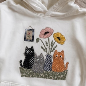May include: An off-white hooded sweatshirt featuring a fabric applique design of three cats sitting in a green floral planter next to a blue patterned vase with pink and yellow flowers, plus a small framed flower picture hanging above.