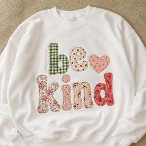 May include: A white crewneck sweatshirt featuring the words "be kind" appliqued on the front. The letters are crafted from various fabrics with green gingham, floral, and polka dot patterns, accompanied by a small pink floral heart shape.