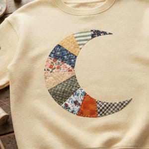 May include: A cream-colored crewneck sweatshirt featuring a large crescent moon applique on the front. The moon is constructed from various fabric patches in floral, striped, checkered, and solid patterns in shades of blue, orange, green, and yellow.