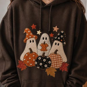 May include: A dark brown hooded sweatshirt featuring a central applique design of three white ghosts. The ghosts wear patterned hats and hold pumpkins with orange, black, and white floral or checkered fabric textures, accented by small stars and leaves.