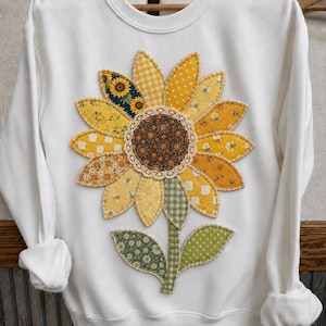 May include: A white long-sleeve sweatshirt featuring a large, central sunflower appliqué. The flower petals are crafted from various yellow and gold patterned fabrics, while the center is dark brown with a cream-colored lace trim border.
