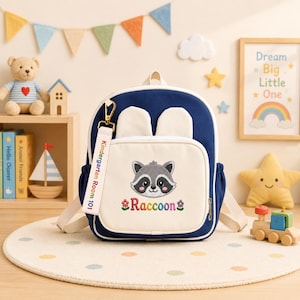 May include: A navy blue and cream backpack featuring a central embroidered raccoon face, the text "Raccoon" with flower icons, and a white fabric keychain tag printed with "Kindergarten Room 101". The bag has two small rounded ears on top.