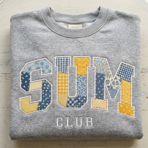 May include: A heather gray crewneck sweatshirt featuring the word SUM in large, patchwork-style letters made from blue and yellow floral and gingham fabric, with the word CLUB embroidered in white thread underneath the large letters.