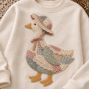 May include: A cream-colored crewneck sweatshirt featuring a large, central applique of a duck wearing a bonnet. The duck is constructed from various fabric patches in muted floral, gingham, and plaid patterns, accented with delicate lace trim.