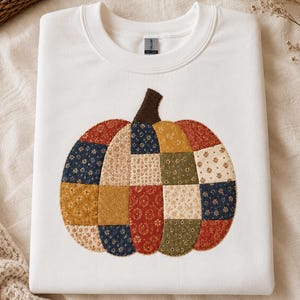 May include: A white crewneck sweatshirt featuring a large, central pumpkin applique. The pumpkin is constructed from various fabric patches in shades of navy blue, mustard yellow, olive green, and rust red, each decorated with small floral patterns.