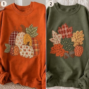 May include: Two crewneck sweatshirts. The first is orange with a quilted-style applique of pumpkins in various fabric patterns. The second is olive green with a quilted-style applique of autumn leaves, a small pumpkin, and two acorns.