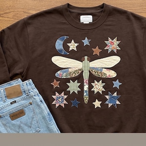 May include: A dark brown crewneck sweatshirt featuring a large central dragonfly appliqué made from various patterned fabric patches, surrounded by smaller star and crescent moon fabric appliqués, paired with light blue denim jeans.
