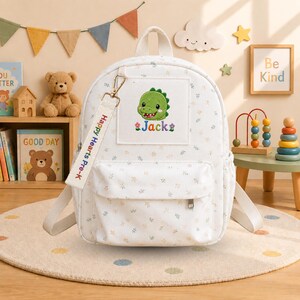 K&ouml;nnte beinhalten: Ein wei&szlig;er Rucksack mit kleinem Blumenmuster, der einen mittigen quadratischen Aufn&auml;her mit einem gestickten gr&uuml;nen Dinosaurier und dem Namen Jack zeigt. Ein wei&szlig;es Schl&uuml;sselband aus Stoff mit dem Aufdruck Happy Hearts Pre-K ist befestigt.