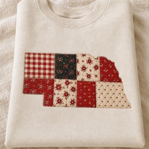May include: A cream-colored crewneck sweatshirt featuring a large, central applique in the shape of the state of Nebraska. The applique is constructed from a patchwork of quilted fabric squares in red, white, and black with floral and gingham patterns.