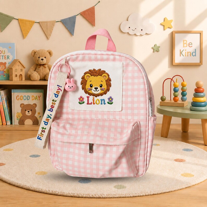 K&ouml;nnte beinhalten: Ein hellrosa-wei&szlig;er Rucksack mit Vichy-Karo-Muster und einem zentralen wei&szlig;en Aufn&auml;her, der einen gestickten L&ouml;wenkopf und darunter den Schriftzug "Lion" zeigt. Ein wei&szlig;es Stoffband am Rei&szlig;verschluss tr&auml;gt den Text "First day, best day!" mit einem kleinen rosa Hasenanh&auml;nger.