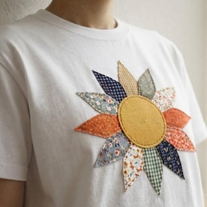 May include: A white short-sleeved t-shirt featuring a large, central sunflower applique. The flower has a solid mustard yellow circular center surrounded by twelve pointed petals, each crafted from different patterned fabrics in shades of blue, orange, green, and floral prints.