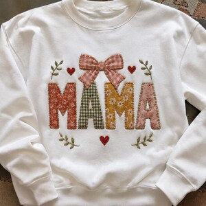 May include: A white crewneck sweatshirt featuring the word MAMA appliqued in various floral and gingham fabric patterns. A pink gingham bow sits above the letters, accented by embroidered red hearts and green leafy sprigs on the front.