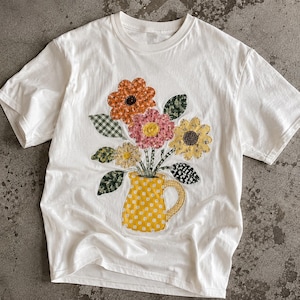 May include: A white short-sleeve t-shirt featuring a central graphic of a yellow polka-dot pitcher filled with a bouquet of multi-colored fabric-style flowers. The flowers include orange, pink, and yellow petals with green patterned leaves.