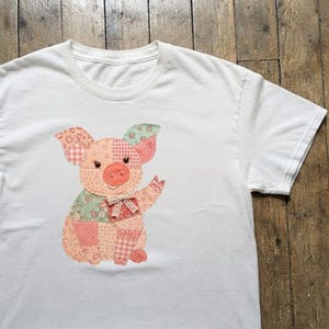 May include: A white short-sleeved t-shirt featuring a central applique of a pig. The pig is constructed from various fabric patches in shades of pink, peach, and sage green with floral and gingham patterns. A small beige lace bow is at the neck.