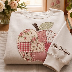 May include: A cream-colored crewneck sweatshirt featuring a large, central apple-shaped patch made of various pink and red floral and gingham fabric squares. A dark brown stem and a green leaf are attached. The text Ms. Evelyn is embroidered on the sleeve.