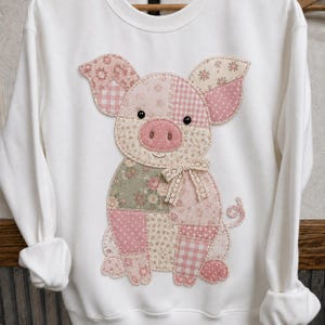 May include: A white long-sleeve sweatshirt featuring a large, central applique of a pig. The pig is constructed from various fabric patches in shades of pink, cream, and sage green, featuring floral, polka dot, and gingham patterns with a lace bow.