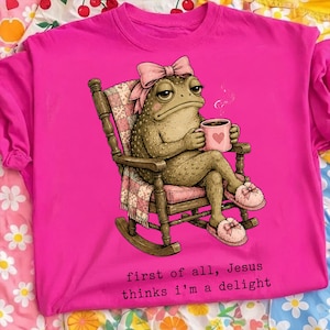 Può includere: Una t-shirt rosa acceso con la grafica di un rospo che indossa un fiocco rosa e pantofole, seduto su una sedia a dondolo in legno mentre tiene in mano una tazza con un cuore. Il testo sottostante recita: first of all, Jesus thinks i'm a delight.