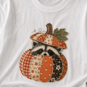 May include: A white t-shirt featuring a central applique design of a raccoon peeking out from inside a large, multi-patterned pumpkin. The pumpkin is constructed from various fabric patches in shades of orange, brown, and black with floral and gingham prints.