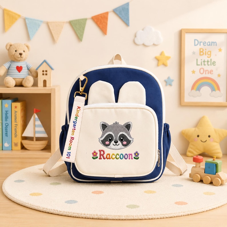 K&ouml;nnte beinhalten: Ein marineblauer und cremefarbener Rucksack mit einem zentralen gestickten Waschb&auml;rengesicht, dem Text "Raccoon" mit Blumensymbolen und einem wei&szlig;en Stoffanh&auml;nger mit der Aufschrift "Kindergarten Room 101". Der Rucksack hat zwei kleine Ohren.