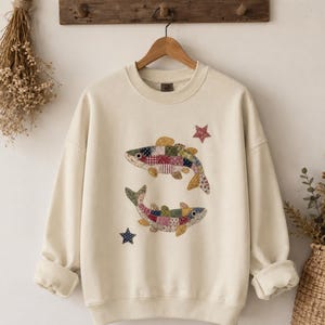 May include: A cream-colored crewneck sweatshirt featuring a central graphic of two fish designed with a colorful patchwork pattern. The fish are surrounded by two small stars, one red and one blue, printed on the front of the garment.