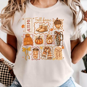 May include: A cream-coloured t-shirt featuring a graphic print with the text "Fall basic" and "AUTUMN". The design includes illustrations of tan boots, an acorn, a beanie, a pumpkin, a candle, a leopard print tumbler, a bow, a coffee cup, and a scarf.