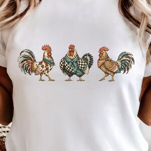 May include: A white t-shirt featuring a graphic print of three roosters standing in a row. Each rooster is illustrated with detailed feathers in shades of brown, red, and teal, and each wears a patterned neck scarf in a different colour scheme.