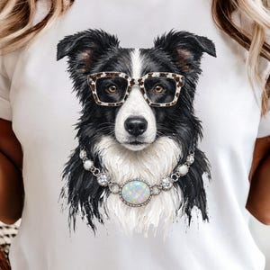 May include: A white t-shirt featuring a graphic print of a black and white border collie dog. The dog is wearing leopard print glasses and a silver-toned necklace with a large, iridescent, oval-shaped central stone and smaller clear crystals.