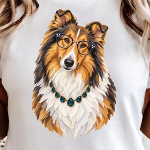 Può includere: Una t-shirt bianca con un'illustrazione dettagliata di un cane collie color zibellino e bianco. Il cane indossa occhiali con montatura blu scuro e una collana dorata con gemme blu a forma di goccia, resa con uno stile pittorico materico.