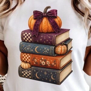 May include: A white t-shirt featuring a graphic print of a stack of four vintage-style books. The top book is purple with a large orange pumpkin and a dark purple bow. The other books are blue and orange, decorated with moons, stars, and flowers.