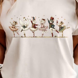 Puede incluir: Una camiseta de color crema con una ilustración gráfica de seis animales antropomórficos vestidos como bailarines de ballet. Los personajes incluyen un ratón, un cisne, un conejo, un gato, otro gato con uniforme de soldado y un ratón con un bastón.