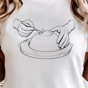 Op de afbeelding: Een wit t-shirt met een zwarte lijntekening van twee handen die een kleikom vormen op een pottenbakkersschijf. Eén hand houdt een klein metalen boetseerwerktuig vast om een detail in de bovenkant te kerven, terwijl de andere hand de zijkant steunt.