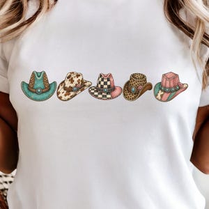May include: A white t-shirt featuring a horizontal graphic print of five distinct cowboy hats. The hats display various patterns including turquoise, cow print, pink and black checkerboard, leopard print, and a pink and turquoise striped design.