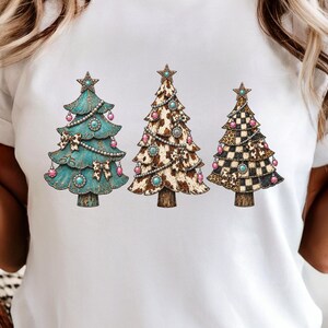 May include: A white t-shirt featuring a graphic print of three stylised Christmas trees. The left tree is teal with bows, the middle tree has a brown and white cow print, and the right tree features a black and white chequered pattern.