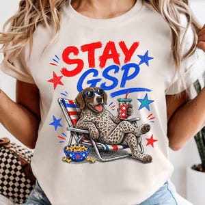 May include: A cream-colored t-shirt featuring a graphic of a German Shorthaired Pointer dog wearing sunglasses, lounging in a beach chair with a drink. The text STAY GSP is printed above in red and blue, surrounded by red and blue star accents.