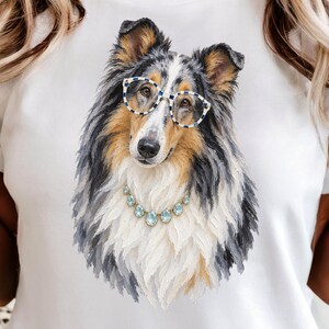 Può includere: Una t-shirt bianca con una grafica dettagliata di un cane collie blue merle. Il cane ha il pelo lungo nei toni del grigio, nero e marrone chiaro, e indossa occhiali a scacchi blu e bianchi insieme a una collana di gemme azzurre a goccia.