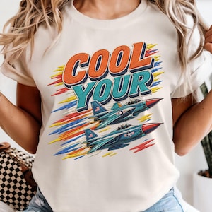 以下が含まれることがあります： クリーム色のTシャツで、オレンジとティールブルーのブロック体で「COOL YOUR」という文字がプリントされています。その下には、2機の青い戦闘機が編隊を組んで飛んでおり、赤、黄、青のスタイリッシュな動きの線が描かれています。