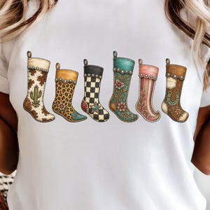May include: A white t-shirt featuring a graphic print of six hanging Christmas stockings. The stockings display various patterns including cow print with a cactus, leopard print, black and white checkerboard with a star, floral teal, pink, and brown leather with a horseshoe.