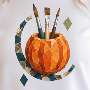 May include: A white t-shirt featuring a graphic of a geometric orange pumpkin-shaped container holding three paintbrushes. The design includes a crescent-shaped mosaic border made of blue, green, and tan triangular tiles surrounding the pumpkin.