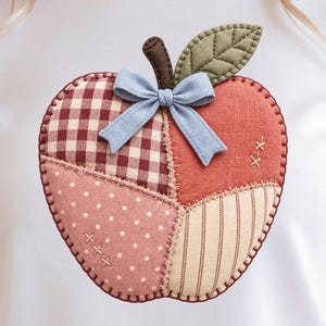 May include: A white t-shirt featuring a large, fabric apple appliqué made of patchwork sections in burgundy gingham, solid rust, pink polka dot, and beige stripe patterns, accented with a brown stem, green leaf, and a light blue ribbon bow.