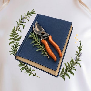 May include: A white t-shirt featuring a graphic of a closed navy blue book with gold-edged pages. Resting on the book are metal pruning shears with orange handles, surrounded by a circular arrangement of green fern fronds and rosemary sprigs.