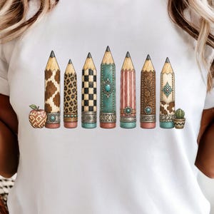 May include: A white t-shirt featuring a graphic print of seven pencils arranged in a row. The pencils display various patterns including cow print, leopard print, checkerboard, and floral designs, accented with turquoise-colored stones and metal bands.
