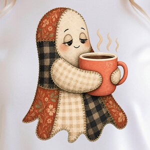 May include: A white t-shirt featuring a graphic of a patchwork ghost holding a steaming orange mug. The ghost is constructed from various fabric patterns including floral prints, black and white buffalo check, and beige gingham with visible stitching.