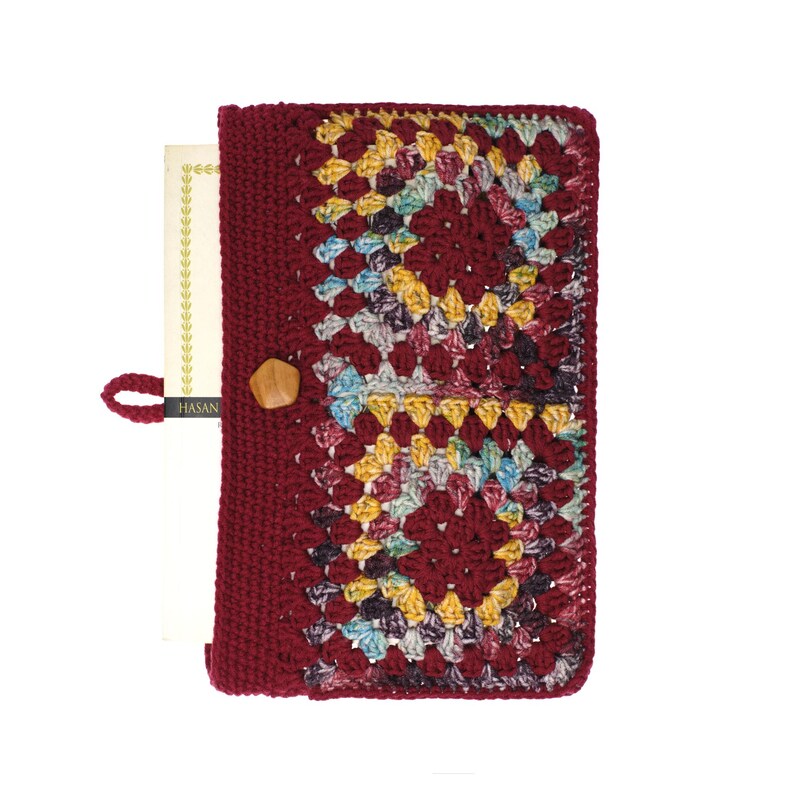 May include: A rectangular book cover made of dark red crochet yarn featuring two large granny square motifs in shades of yellow, blue, and purple. The cover includes a solid dark red crocheted spine, a small wooden pentagonal button, and a loop.