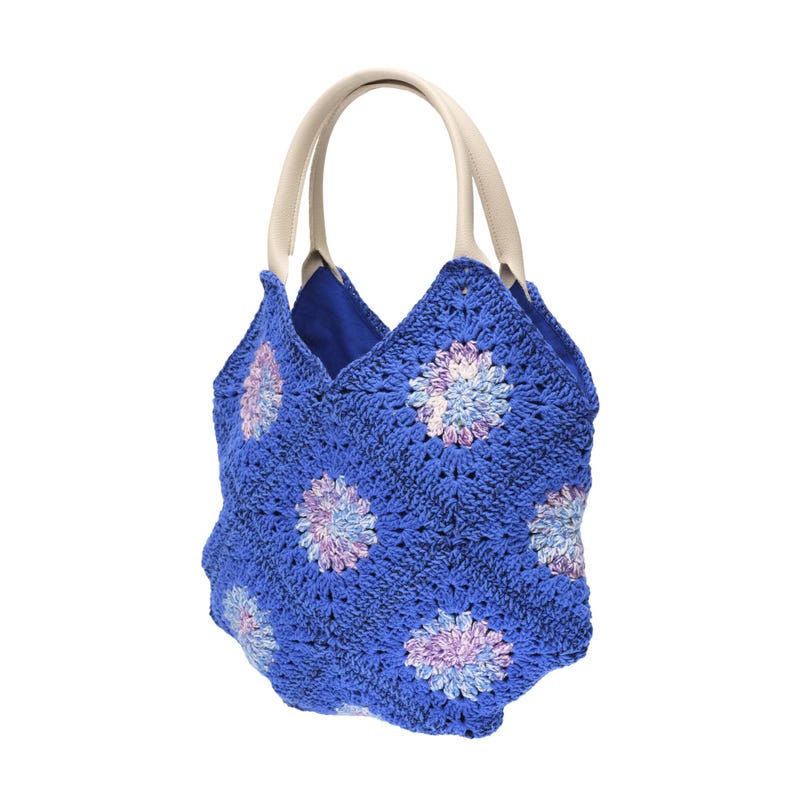 May include: A blue crochet tote bag with a floral pattern in shades of purple, white, and blue. The bag has cream-colored handles and a blue interior. The bag is approximately 12 inches tall.